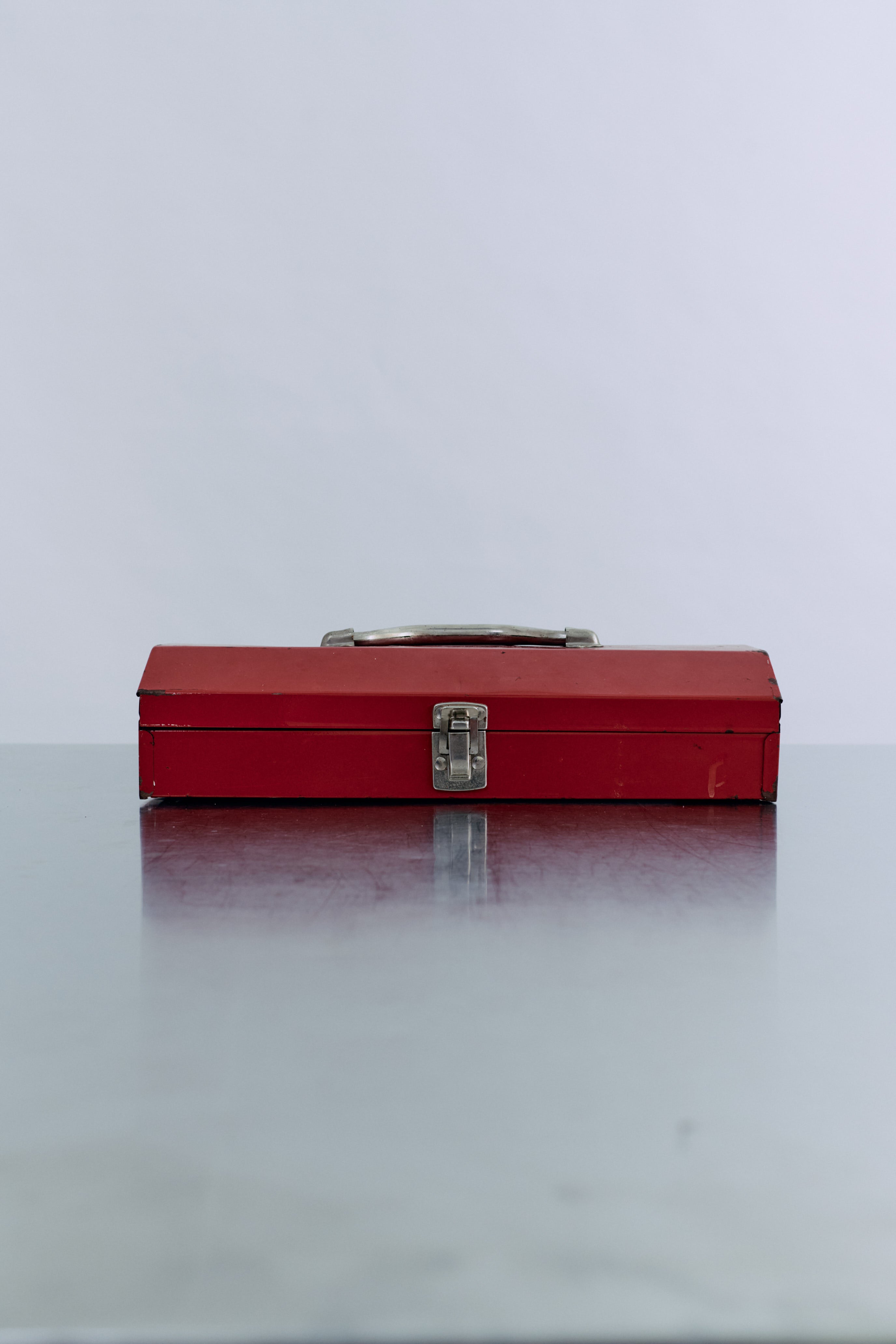 A red metal toolbox with a silver handle, resting on a reflective surface against a light background.