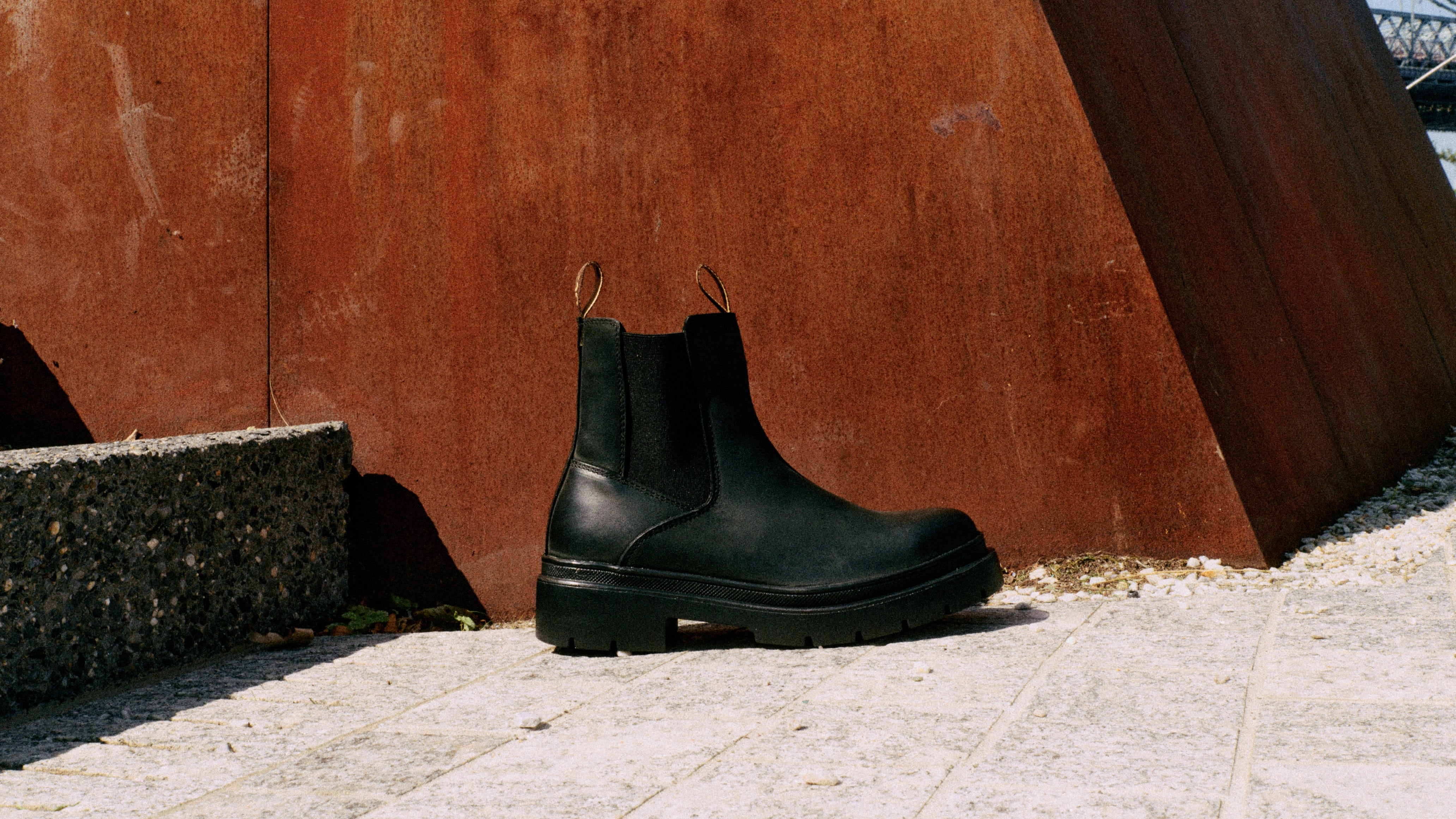 Black Chelsea boots on a stone surface with a rust-colored metal wall in the background.