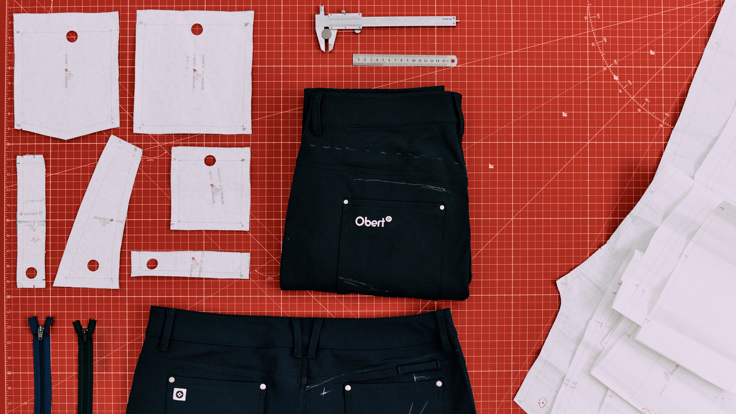 A flat lay of sewing patterns, black pants, zippers, and measuring tools on a red grid background.