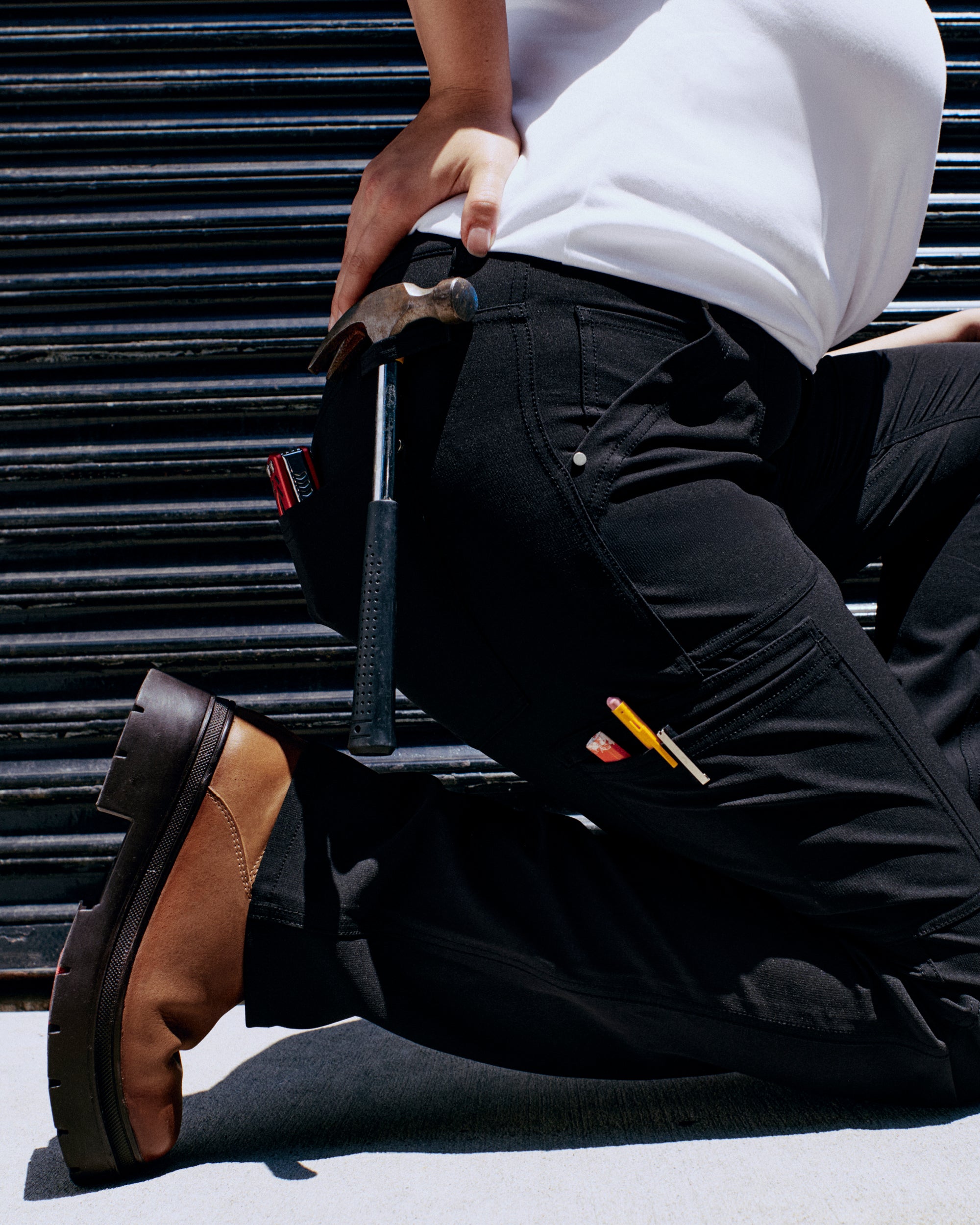 A person kneels, wearing black pants with tools like a hammer and pencils visible in pockets, against a textured background.