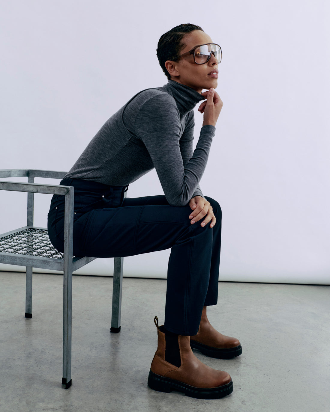 A person sits on a metal chair, wearing a gray turtleneck, dark pants, and brown boots, with large glasses and a thoughtful expression.